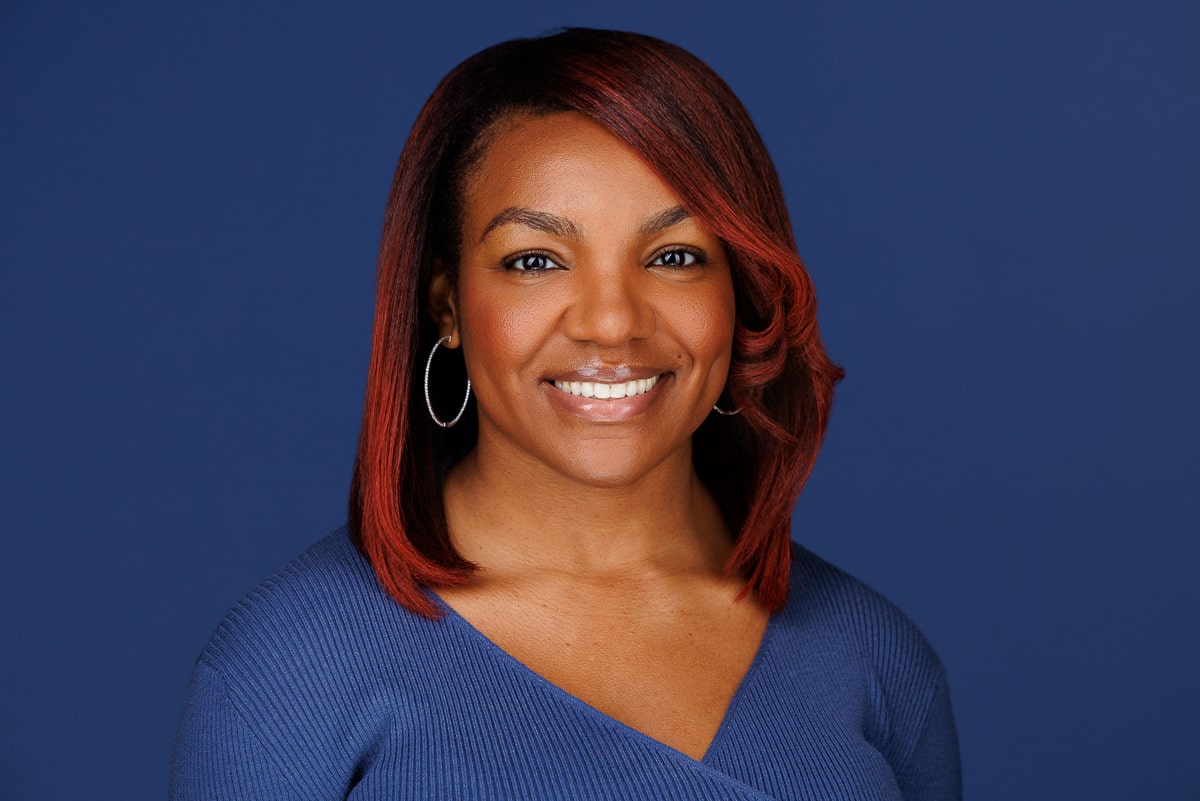 Woman with shoulder-length red hair and hoop earrings, wearing a blue top, smiles in a professional business headshot against a solid blue background.
