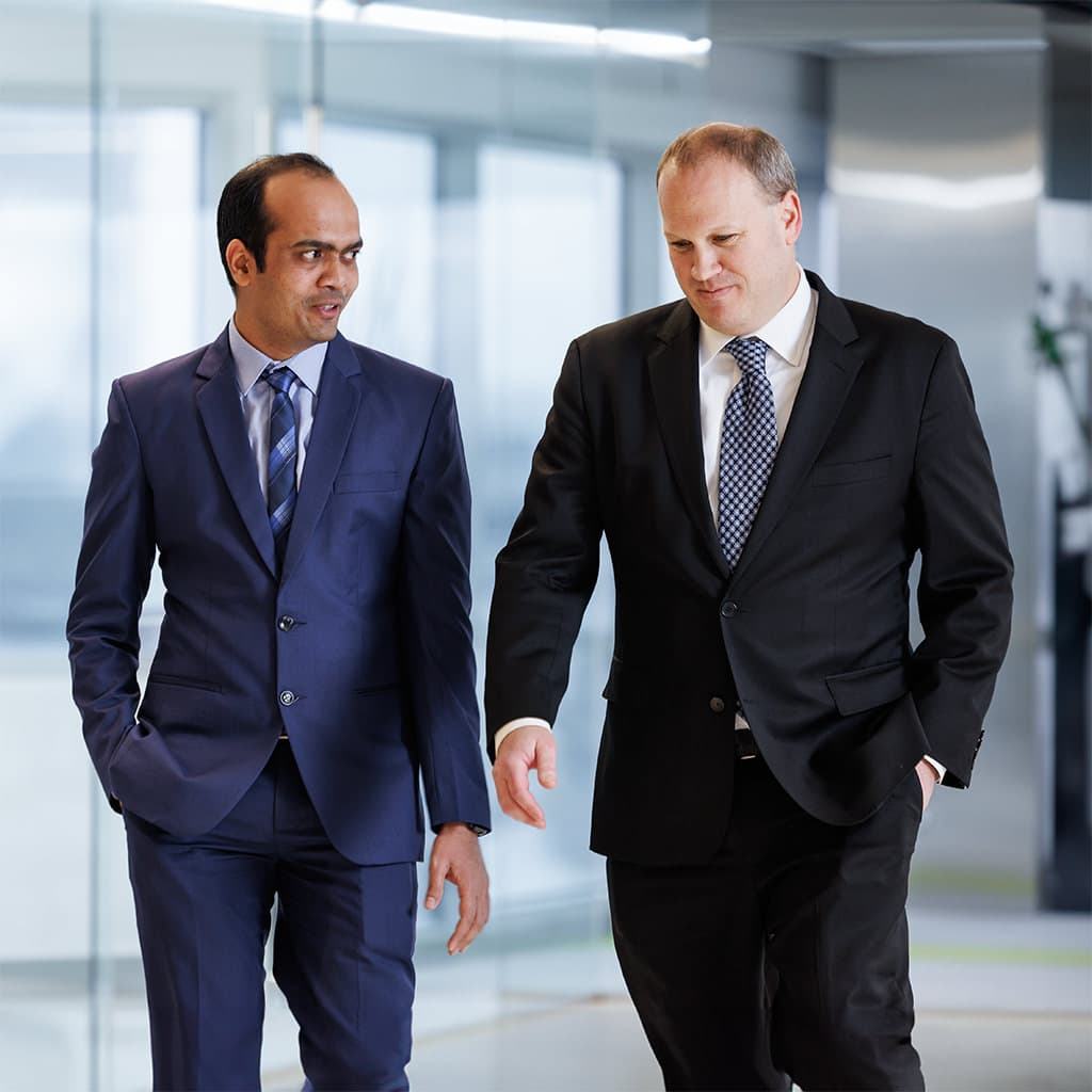 Two men in business suits walking and talking in a modern office hallway with glass walls.