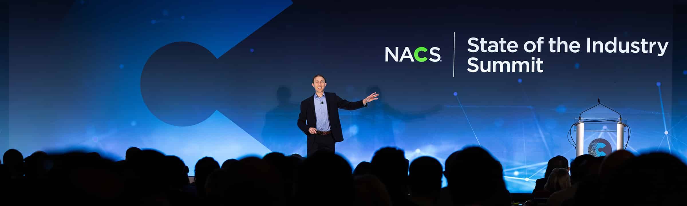 Keynote speaker presenting on stage during a corporate conference as attendees watch from the audience at the NACS State of the Industry Summit.