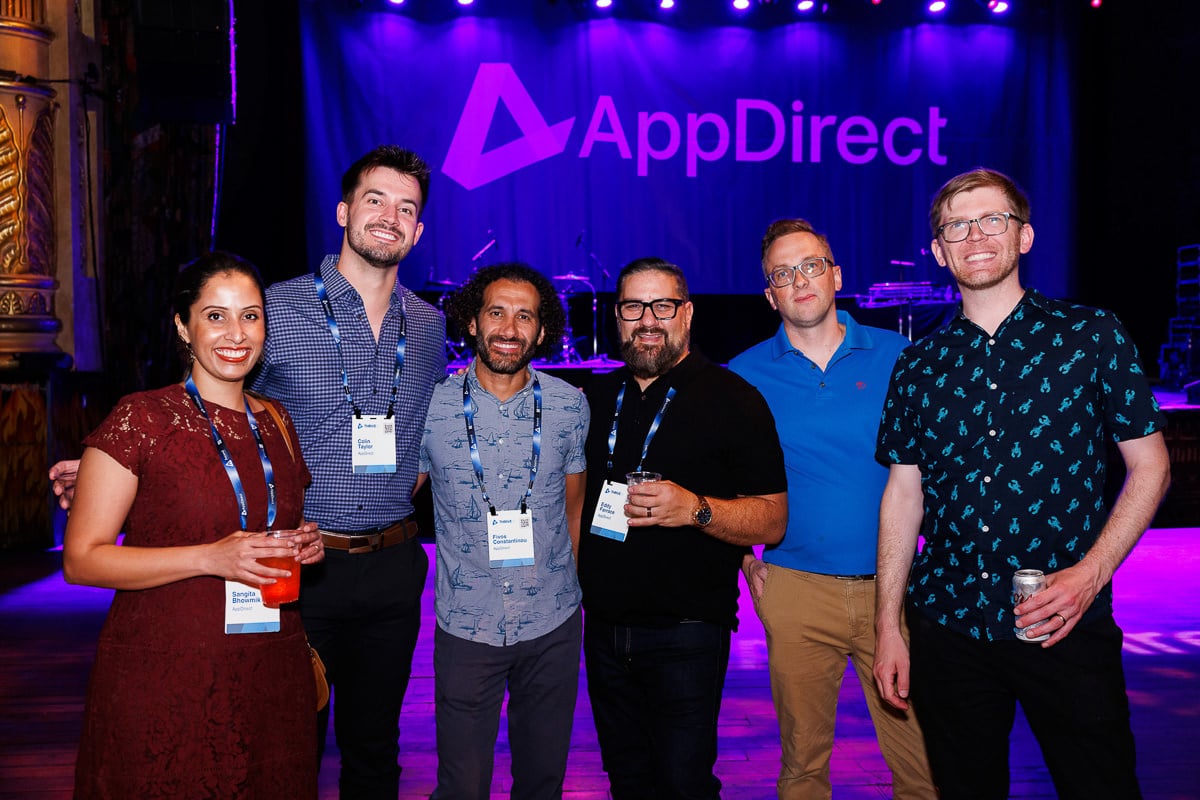 A group of six people standing together in front of a stage with an "AppDirect" backdrop. They are smiling and holding drinks, captured by the Chicago corporate event photography team at Tori Soper Photography.