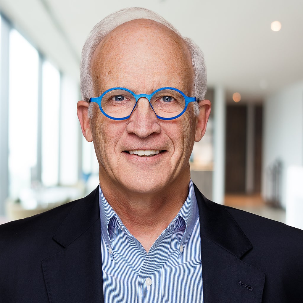 Professional headshot of a senior executive wearing blue glasses, taken in a modern office setting