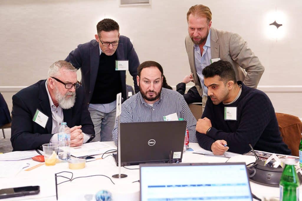 Five men are gathered around a laptop at a table, focusing intently on the screen. Papers and a drink are on the table.