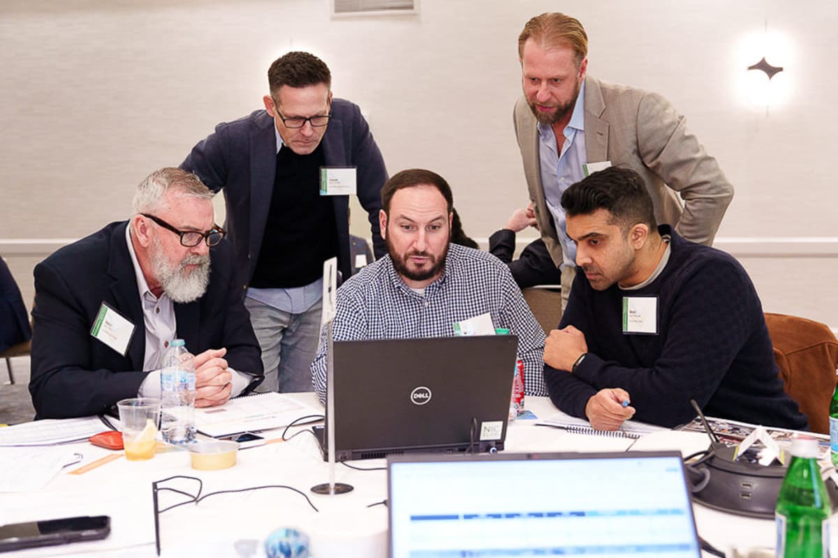 Five men are gathered around a laptop at a table, focusing intently on the screen. Papers and a drink are on the table.