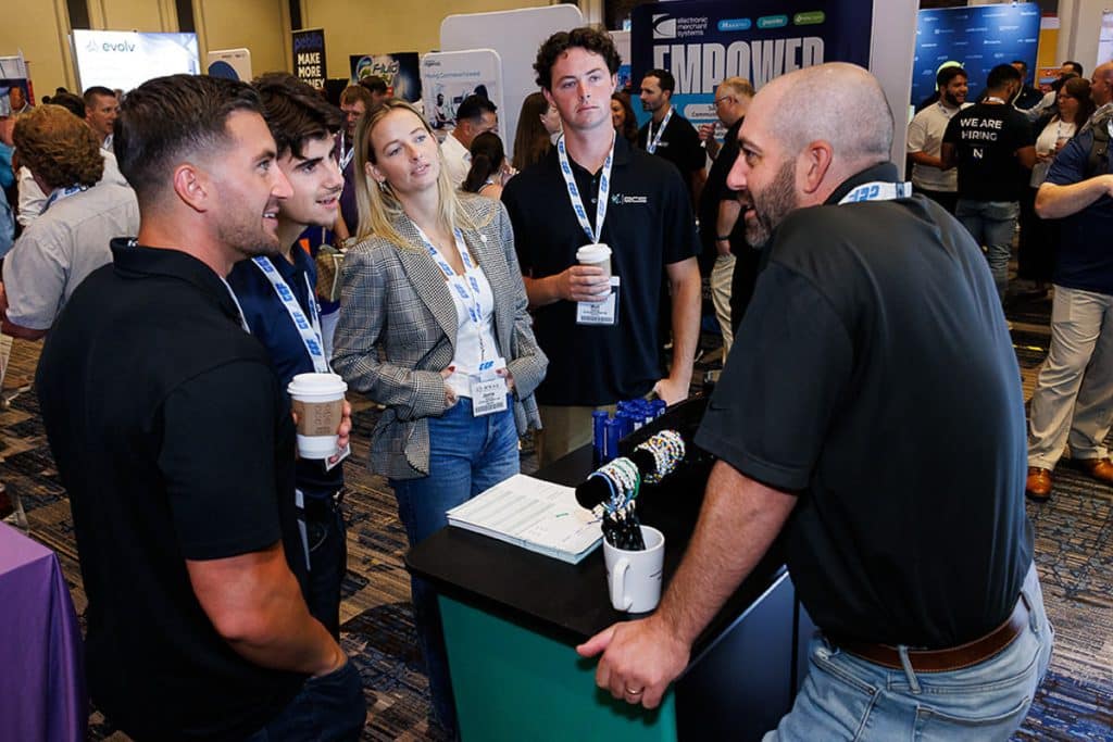 A group of five people stand and converse at a conference booth, holding coffee cups. They are wearing casual business attire with lanyards around their necks.