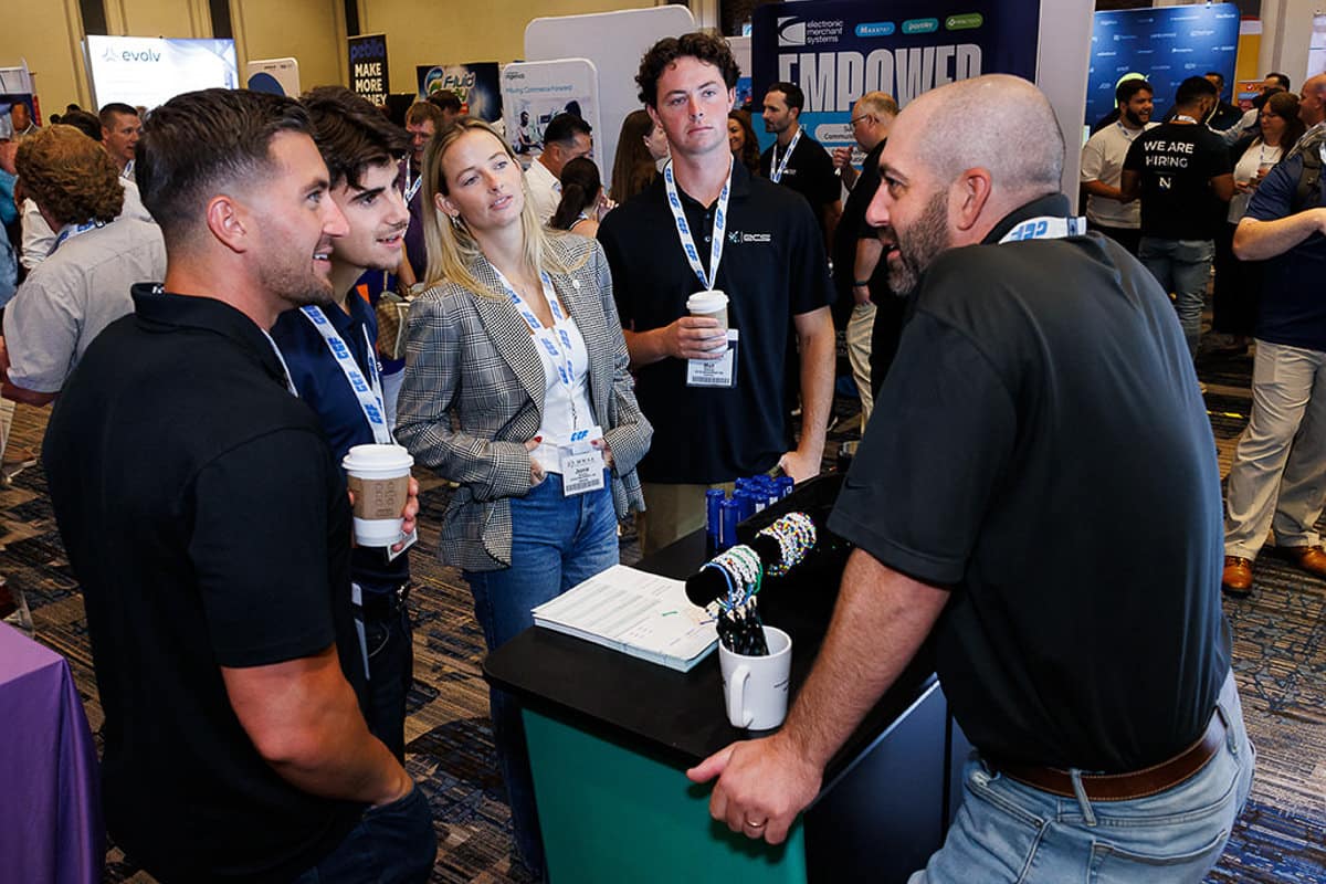 A group of five people stand and converse at a conference booth, holding coffee cups. They are wearing casual business attire with lanyards around their necks.