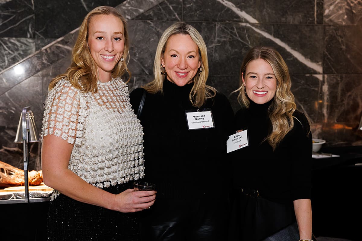 Three smiling women in formal attire stand together in front of a marble wall at an indoor event.