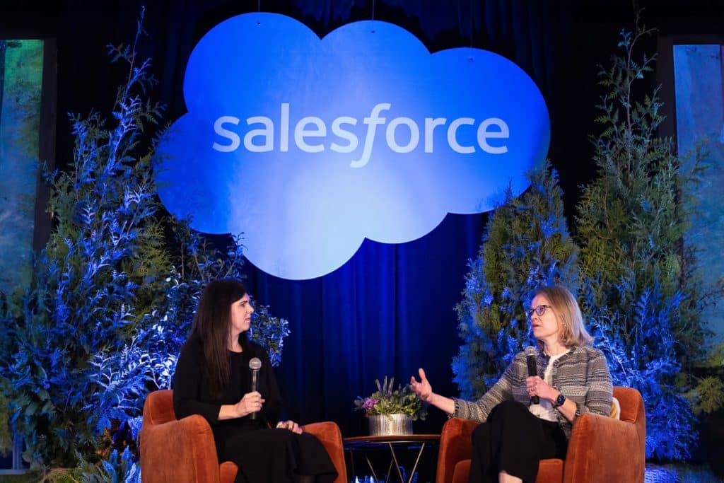 Two women seated on stage with microphones, engaged in conversation. A large illuminated "Salesforce" logo is displayed above, with greenery in the background.