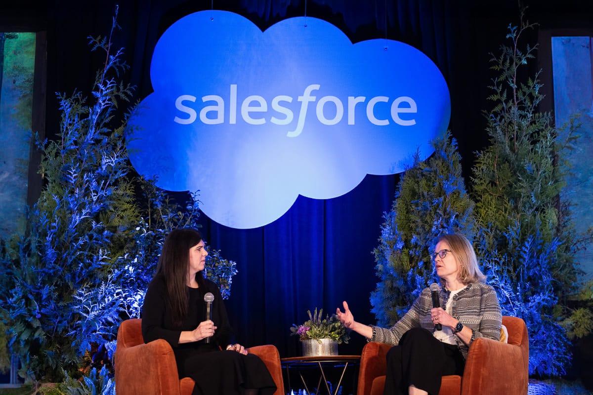 Two women seated on stage with microphones, engaged in conversation. A large illuminated "Salesforce" logo is displayed above, with greenery in the background.