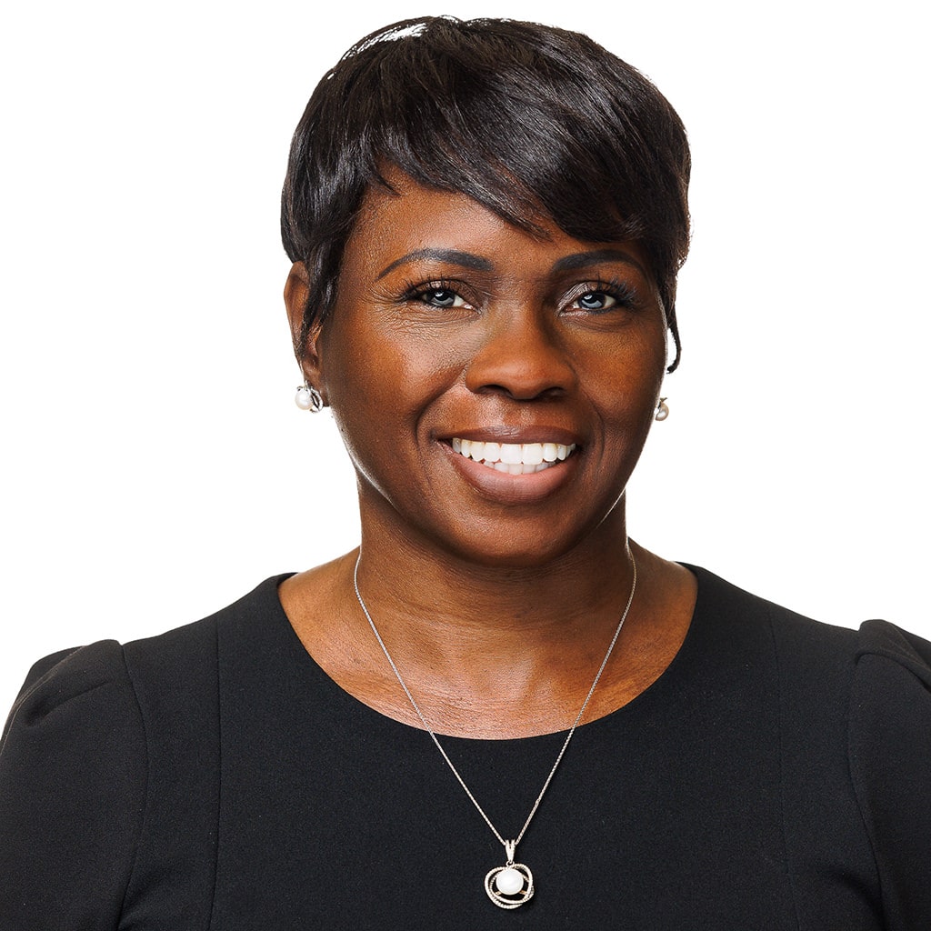 A person with short dark hair smiles, wearing a black top and a necklace with a circular pendant, against a plain white background.