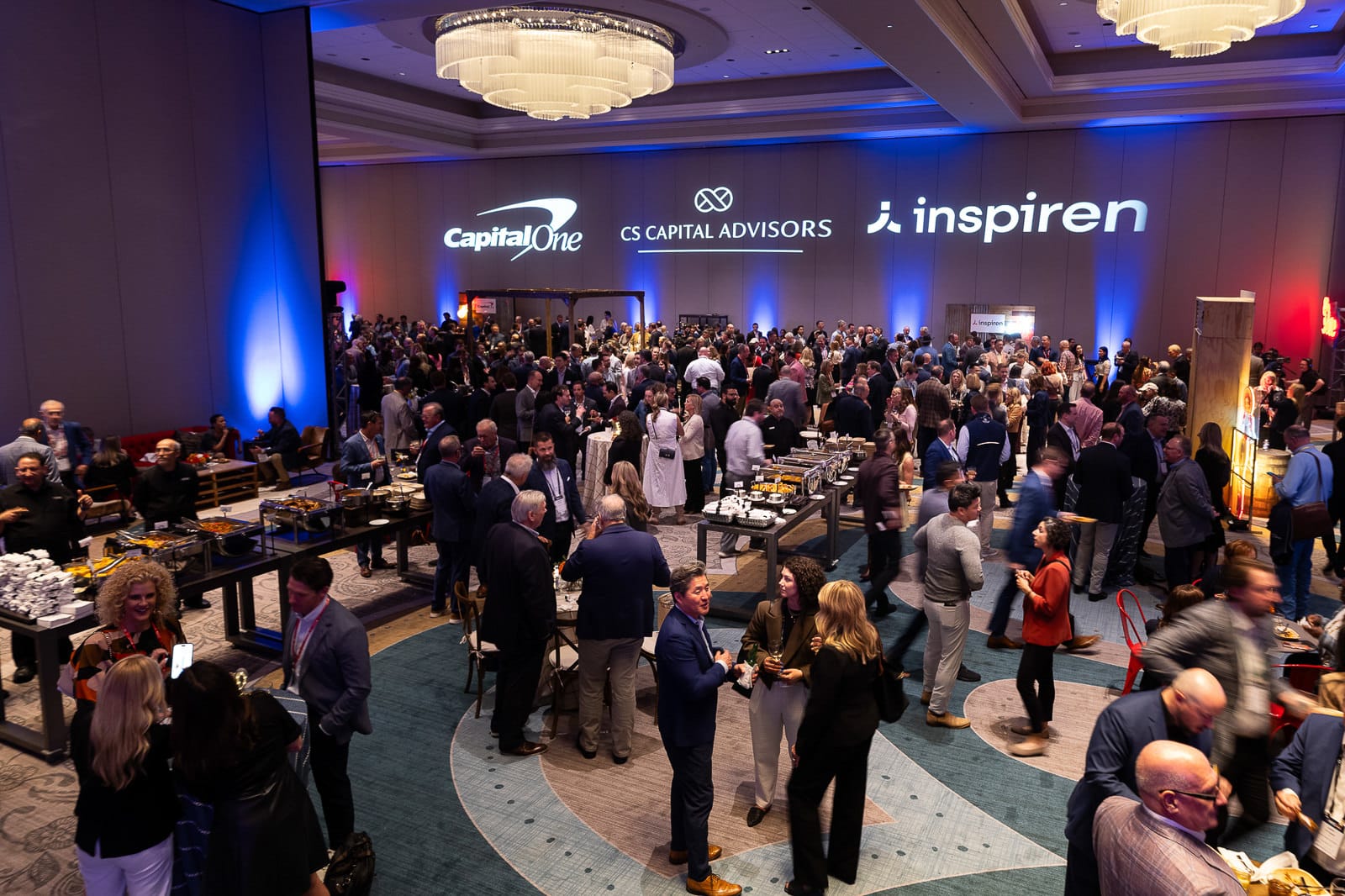 Large hotel ballroom reception with hundreds of attendees mingling around buffet tables; sponsor logos projected on the back wall.