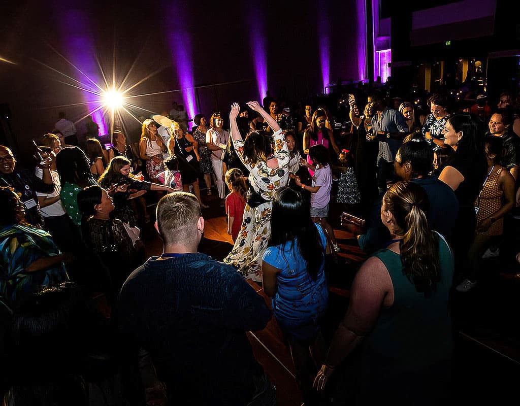 A group of people dancing and socializing at a party. The room is dimly lit with purple lights, and a bright flash is visible in the background.