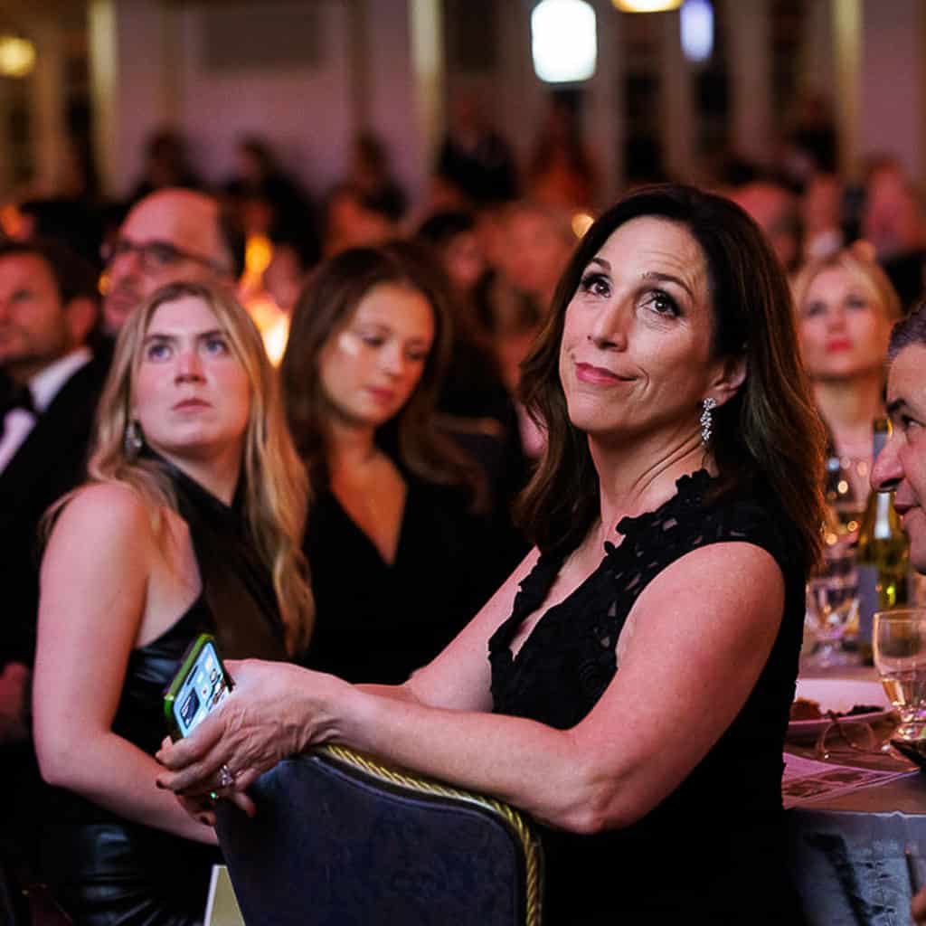 A woman in a black dress sits at a table, holding a phone, surrounded by others in formal attire at an indoor event.